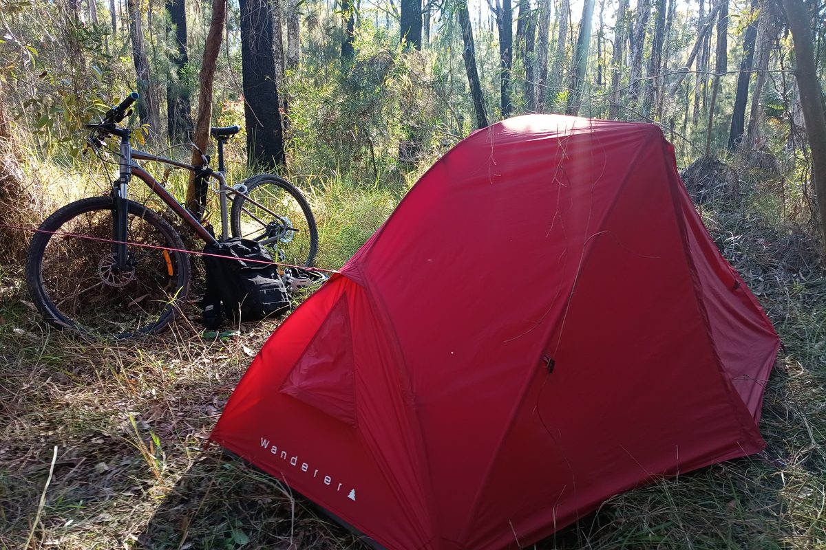 Illustrative photo of a loaded bicycle parked beside a small red backpacking tent in a dry forest