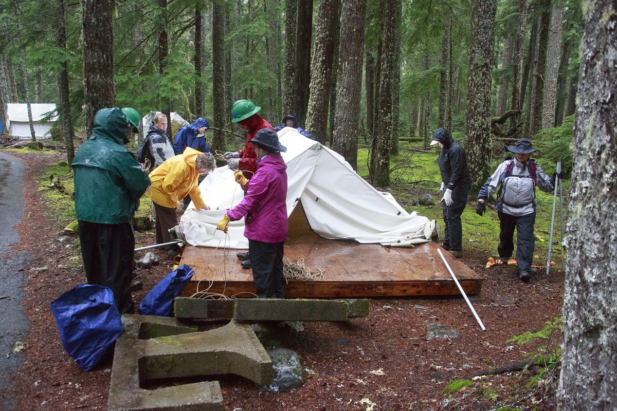 Illustrative photo of a group of volunteers taking down a canvas tent on a wooden platform at a forest campground