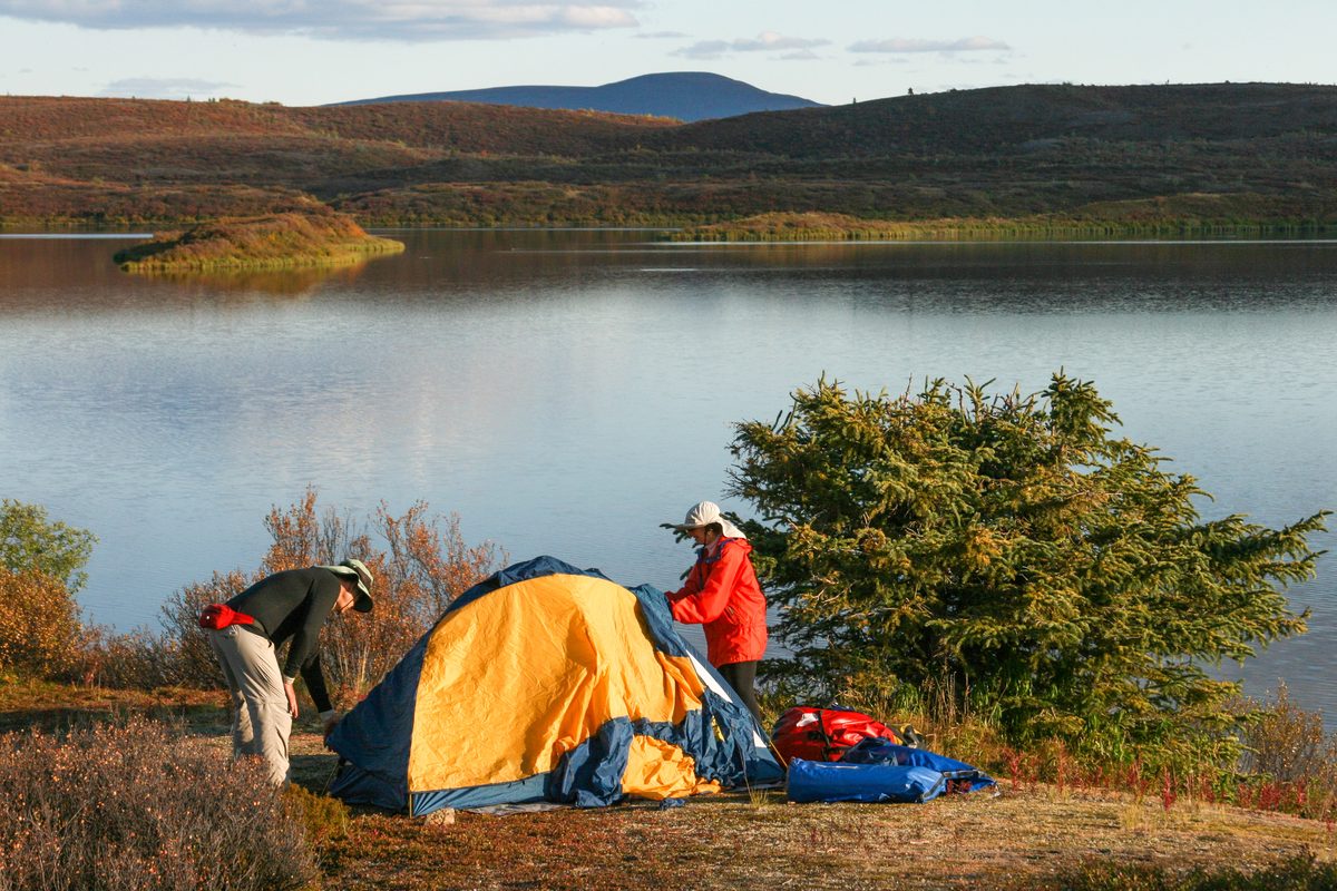 Illustrative photo of two campers pitching a tent beside a lake on an autumn evening