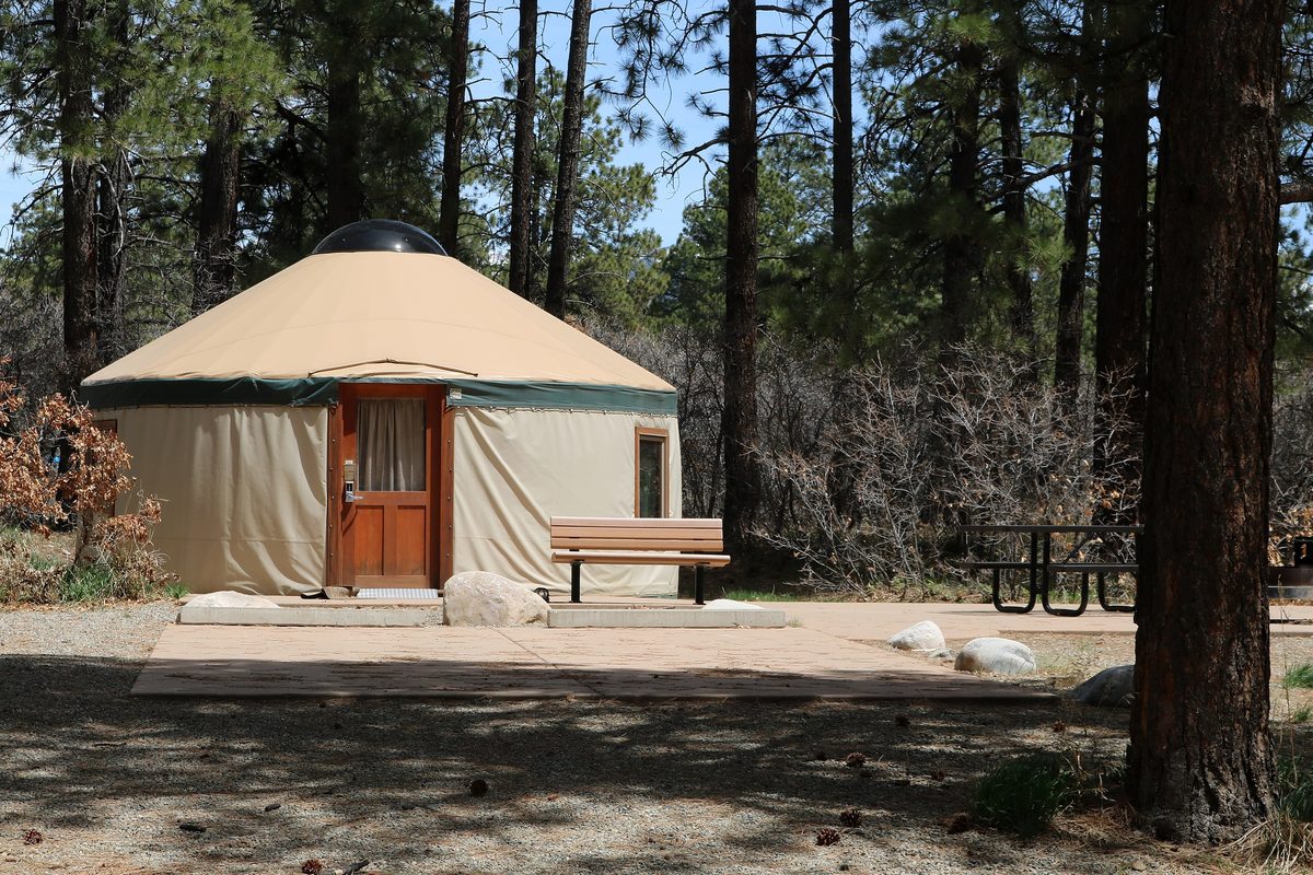 Illustrative photo of a canvas yurt with a wooden door and deck bench at a forested state park campsite