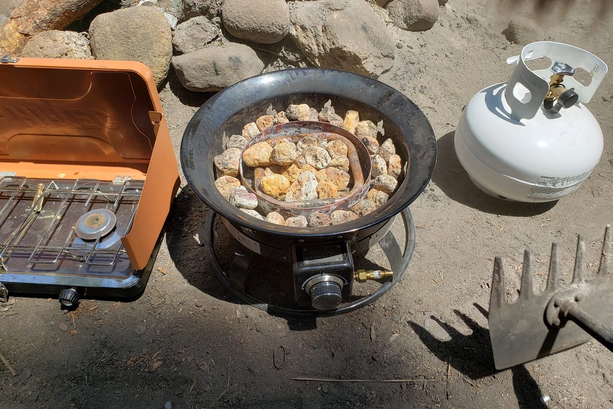 Illustrative photo of a two-burner camp stove, a gas fire pit and a propane cylinder outdoors on sandy ground