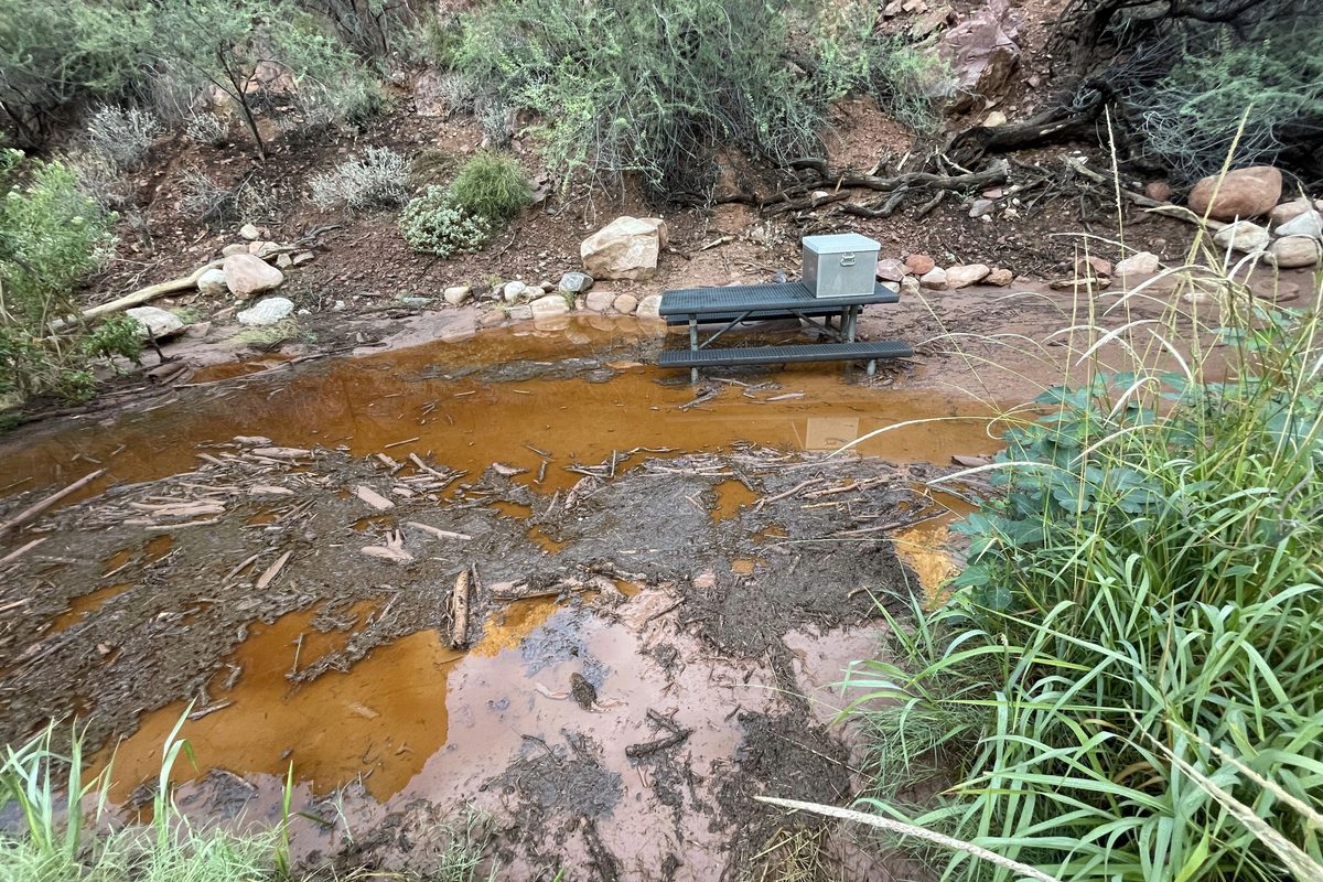 Illustrative photo of a canyon campsite with a picnic table and food box surrounded by mud, debris and standing water after a flash flood