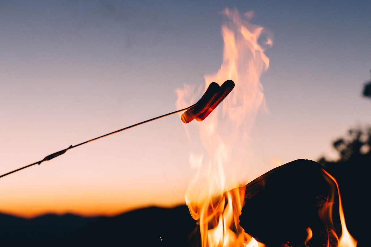 Hot dog cooking on a stick over a campfire at dusk