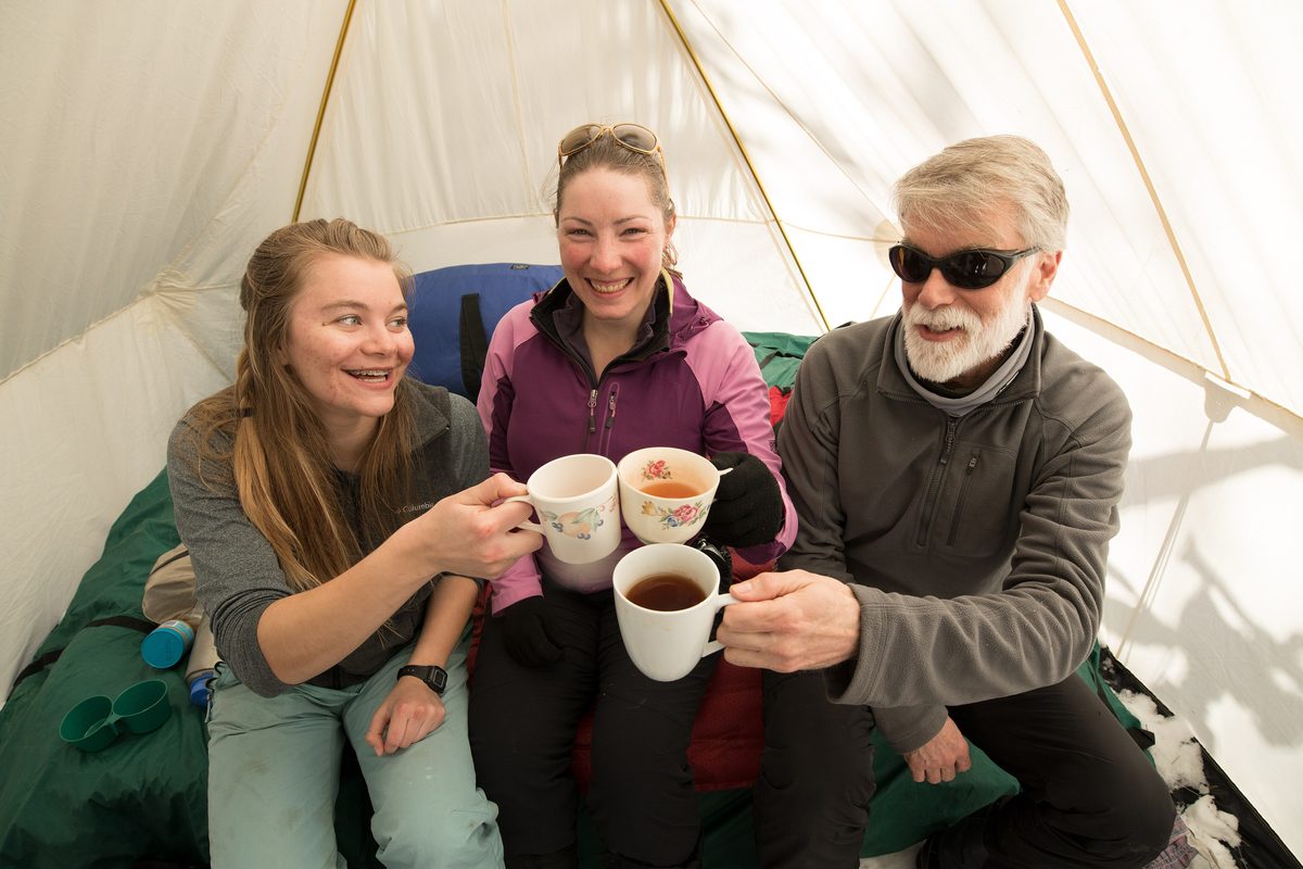 Illustrative photo of three campers in warm layers sharing hot drinks inside a canvas tent on a winter trip